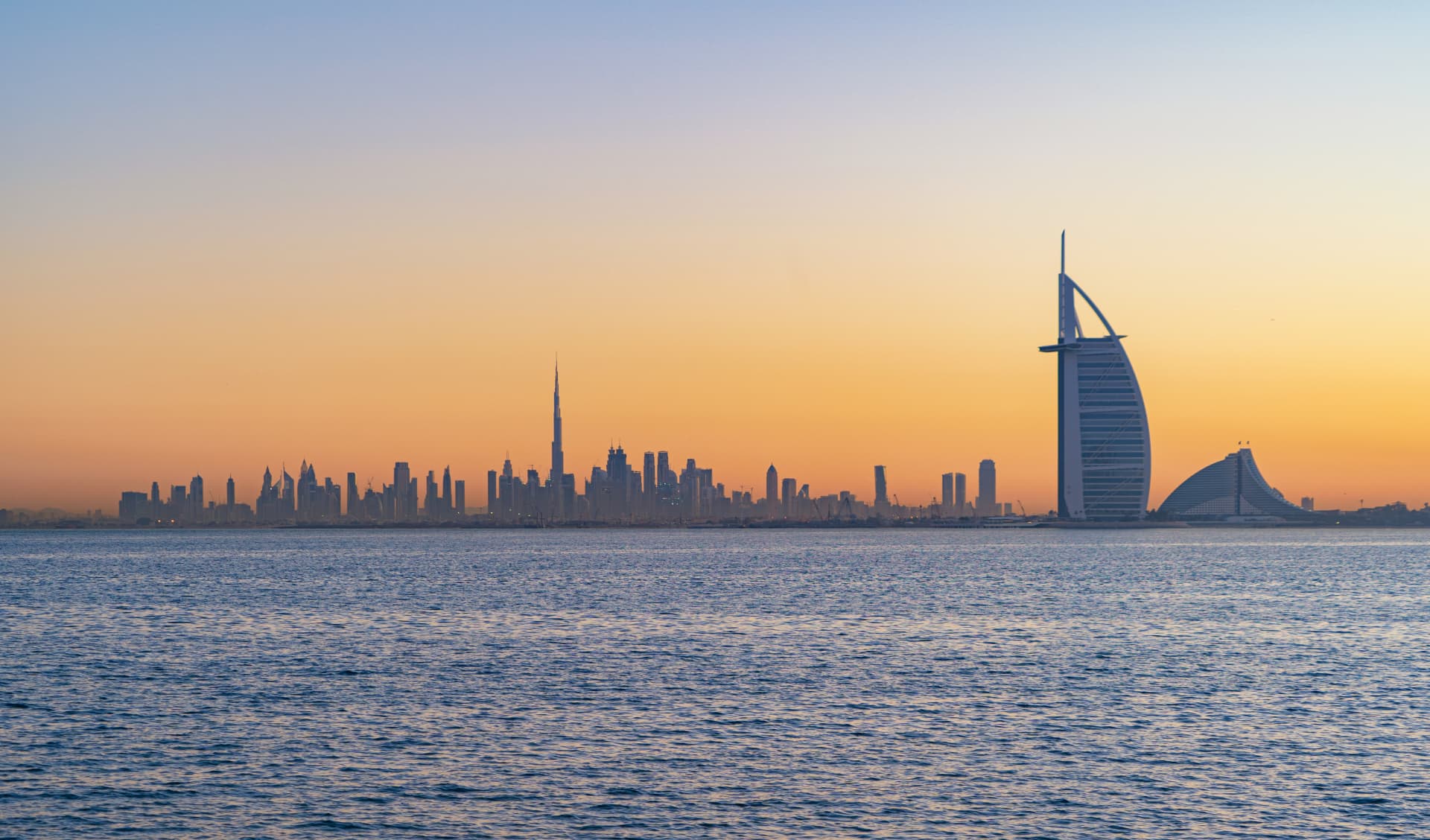 Dubai skyline at sunset seen across the water, with the Burj Khalifa and Burj Al Arab on the horizon